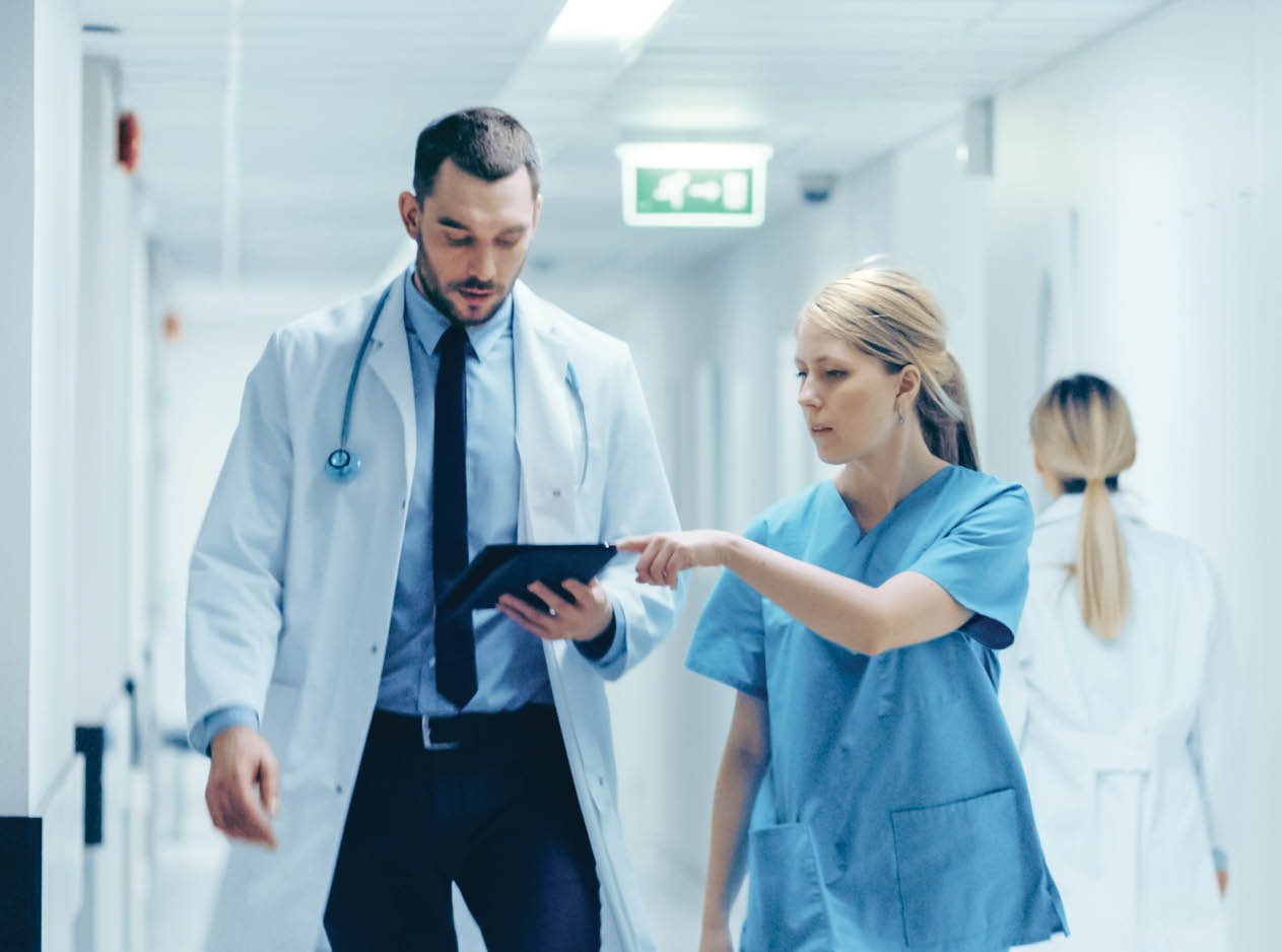 Female Surgeon and Doctor Walk Through Hospital Hallway, They Consult Digital Tablet Computer while Talking about Patient's Health. Modern Bright Hospital with Professional Staff.